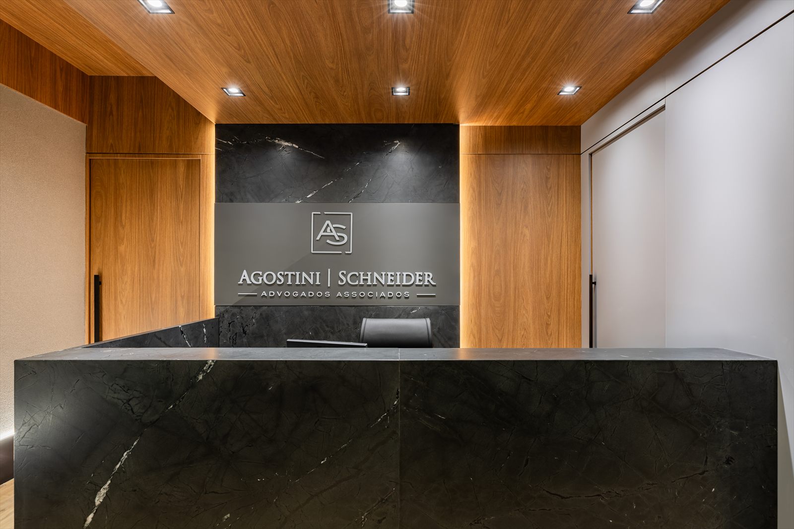 Recepção do escritório Agostini | Schneider, com balcão em pedra e painel de madeira.