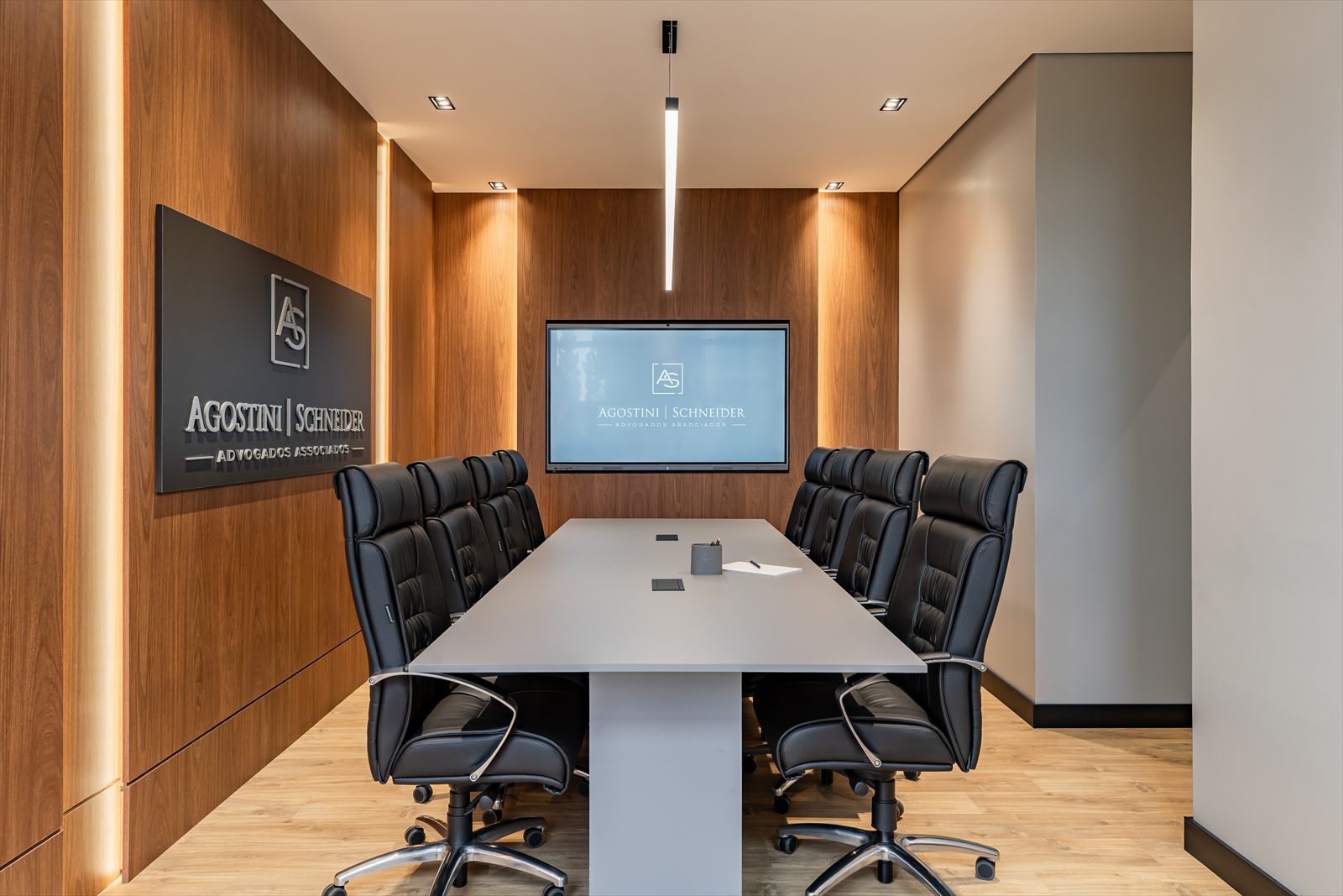 Sala de reuniões do escritório, com mesa central, cadeiras pretas e painel de madeira.